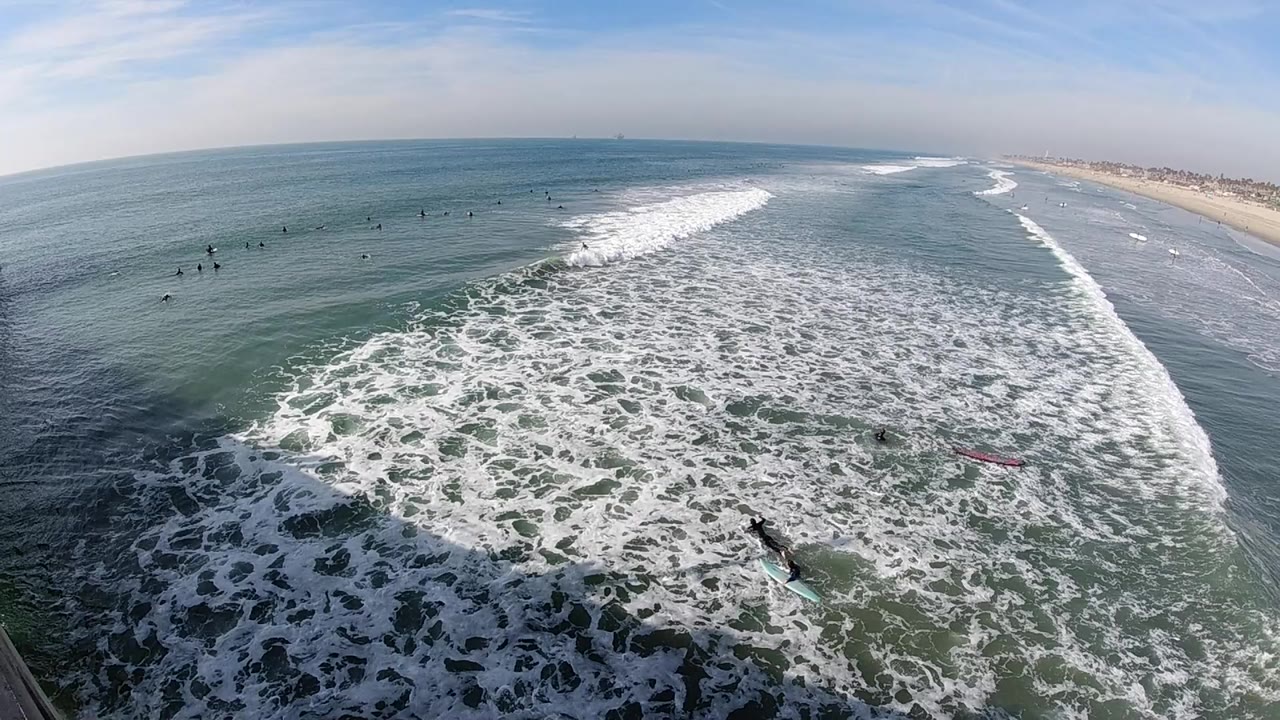 Elevated Huntington Beach ocean view with a broad band of white surf in the center, open water at left, sandy coastline at right, and distant water users appearing as small dark marks.