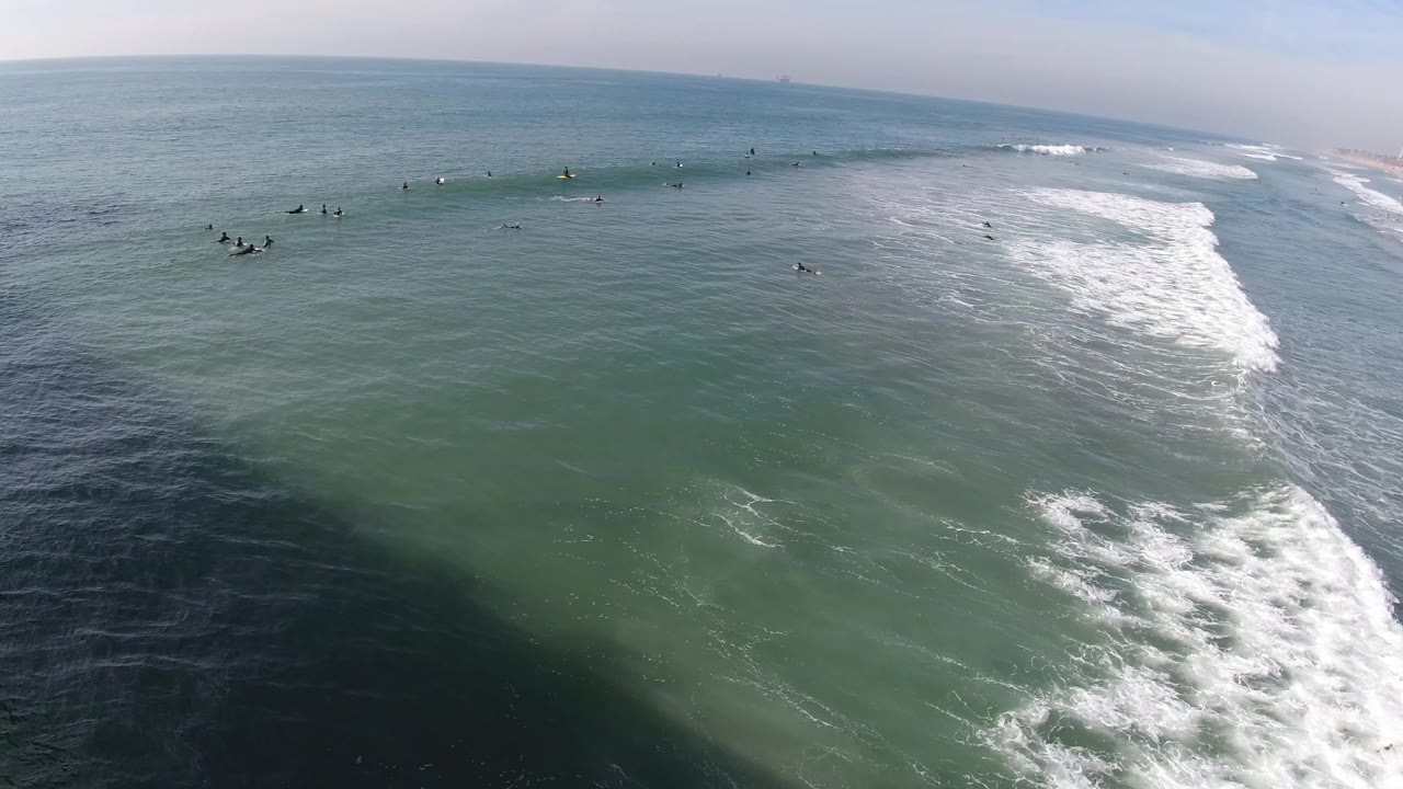 Aerial view over Huntington Beach water with scattered surfers near a white breaking wave line and the sandy shoreline faintly visible to the right.
