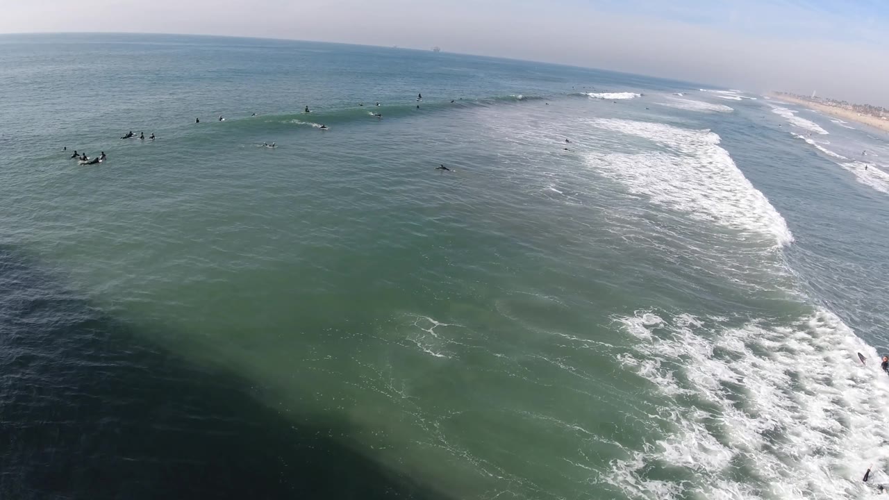 Aerial view of Huntington Beach surf with surfers spaced across the upper water, white foam bands moving diagonally, and the shoreline extending along the far right.
