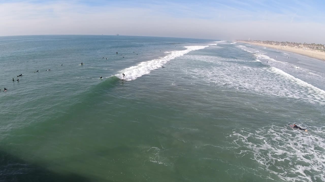 Aerial frame from Huntington Beach with the rider lower on the face of the wave, a broad white foam band cutting across the water, and the beach receding into haze.