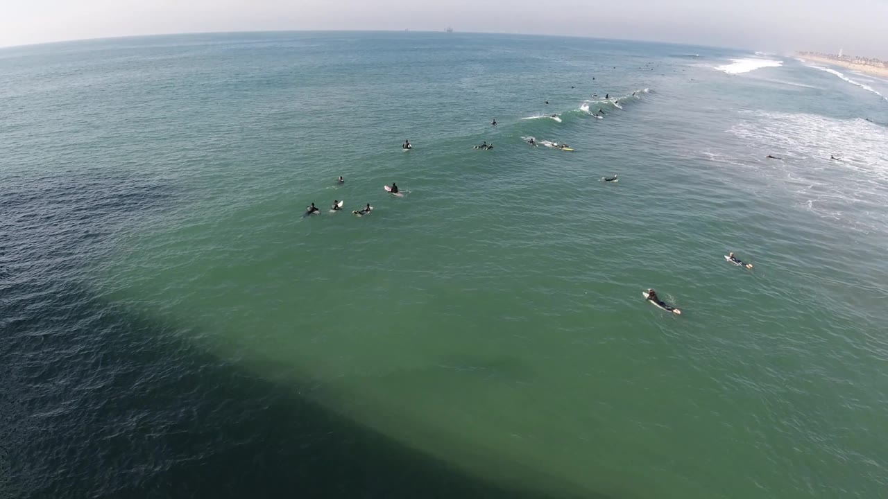 Aerial Huntington Beach view with a diagonal band of breaking surf on the right, multiple surfers waiting offshore, and a hazy coast in the background.
