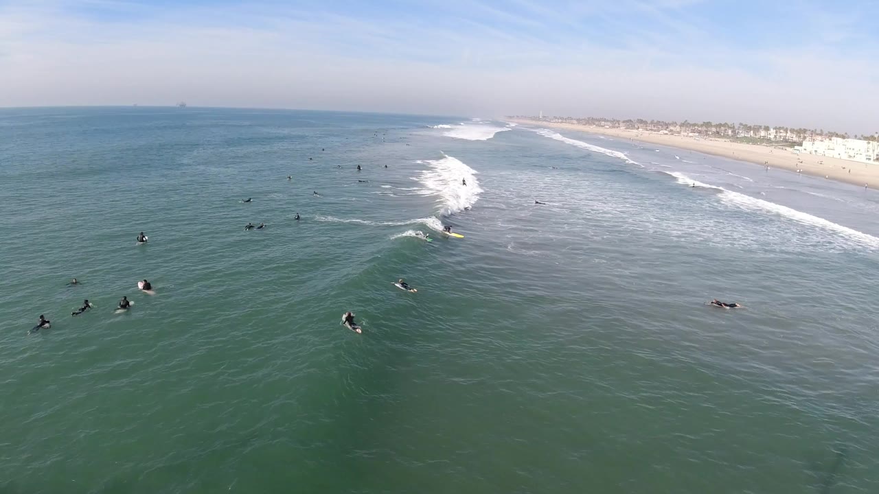 Aerial view over Huntington Beach, California, with scattered surfers in green-blue water and a white wave breaking diagonally toward the sandy shoreline.