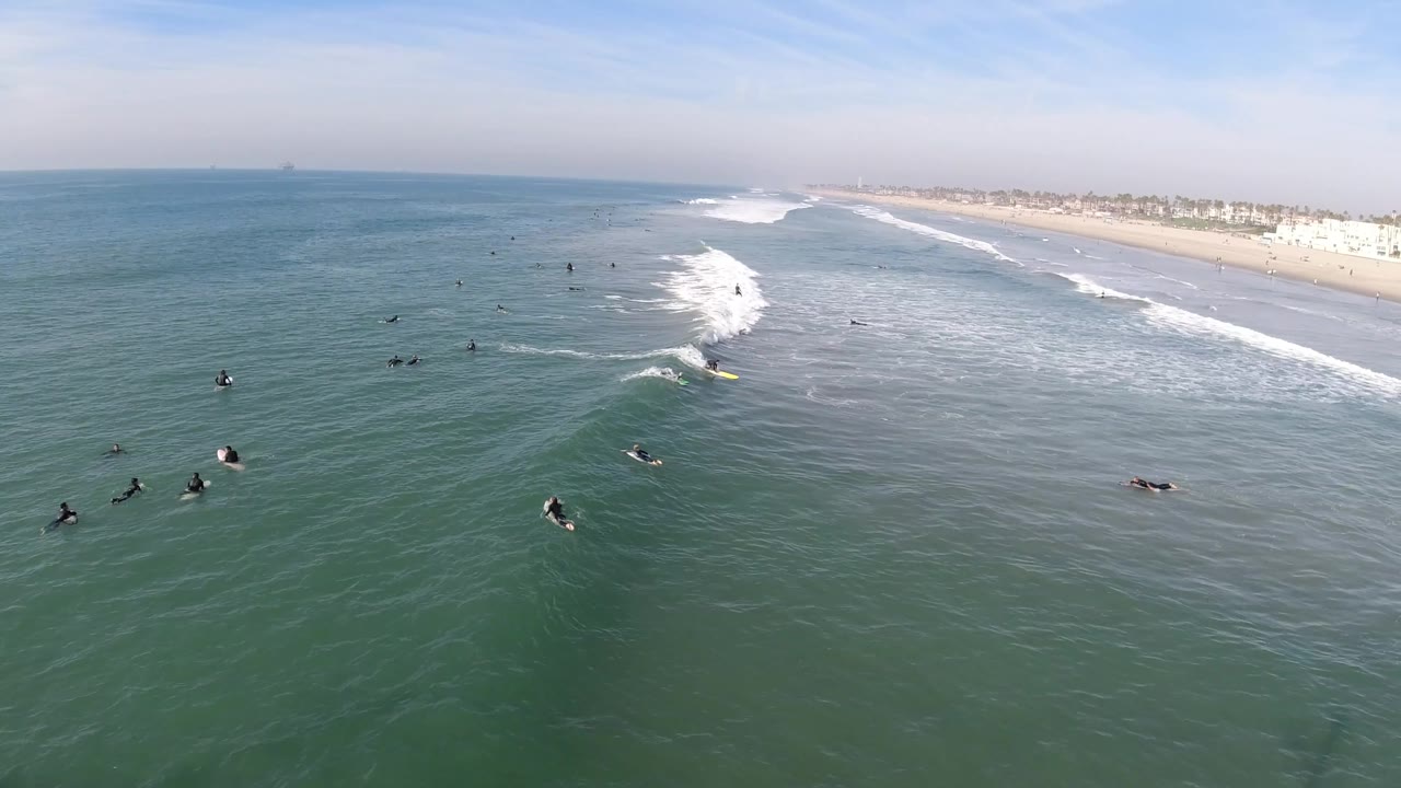 Surfers sit offshore as a white wave line approaches the beach.