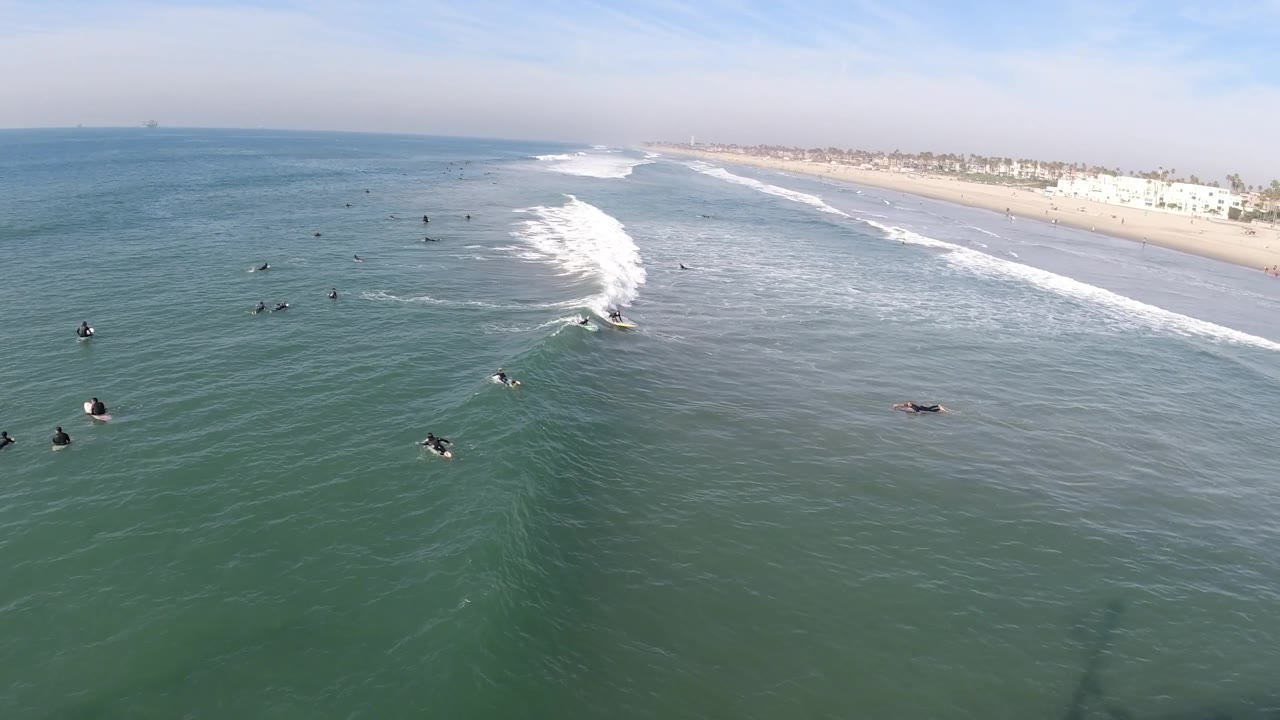Wide aerial view of Huntington Beach with surfers in the water, a broad whitewater section curling toward shore, and a hazy line of beach buildings beyond the sand.