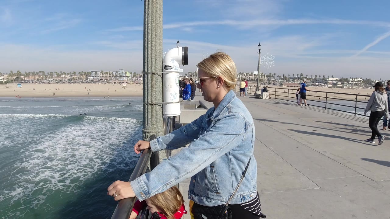 The woman and child remain beside the Huntington Beach pier railing as a wave line moves across the water and pedestrians continue along the walkway behind them.