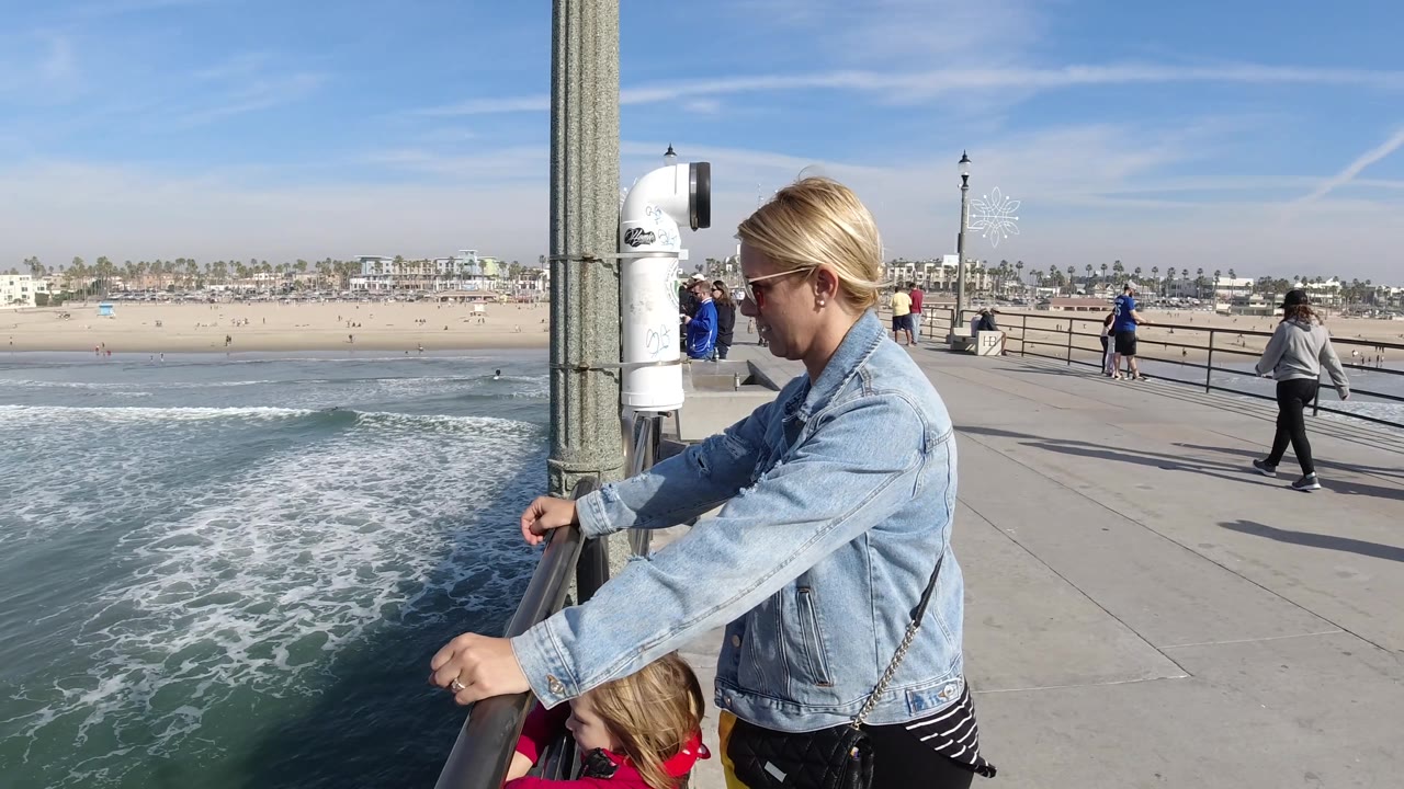 A closer right-side view shows the woman leaning on the railing above the child, with Huntington Beach surf, beachgoers, and a clear sky stretching across the background.
