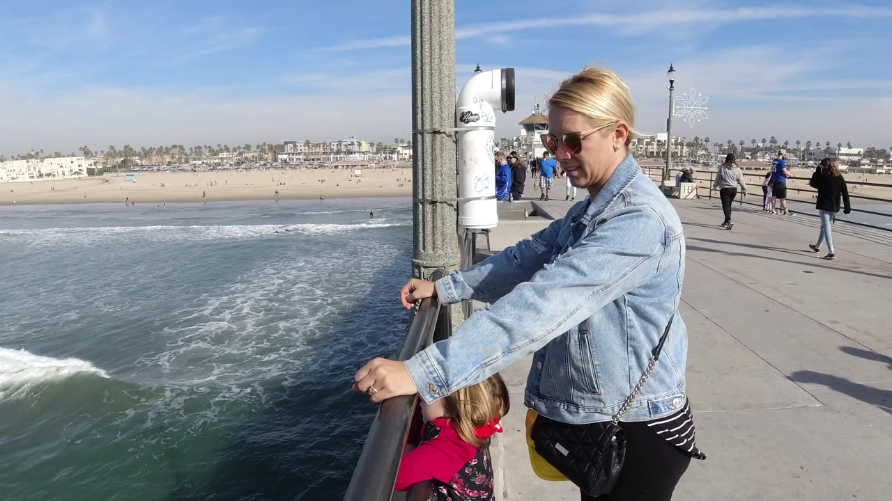 At Huntington Beach, a woman in sunglasses and a denim jacket stands with a child at a pier railing while waves roll below and the sandy beach stretches behind them.