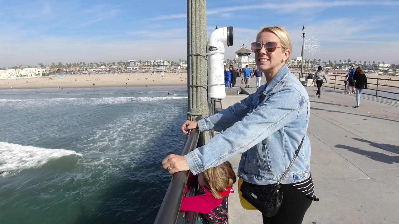 The woman turns toward the camera with a smile while holding the pier rail, the child remains partly visible below, and the beach, surf, and walkway pedestrians fill the background.
