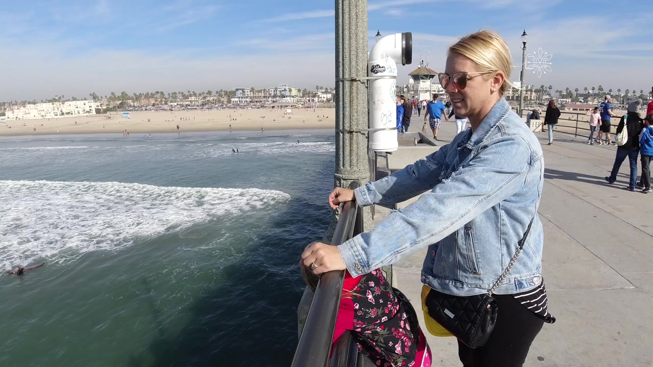 At 0:01 in Huntington Beach, California, the woman remains at the railing near a child in pink as white surf spreads across the water and pedestrians move along the pier walkway.