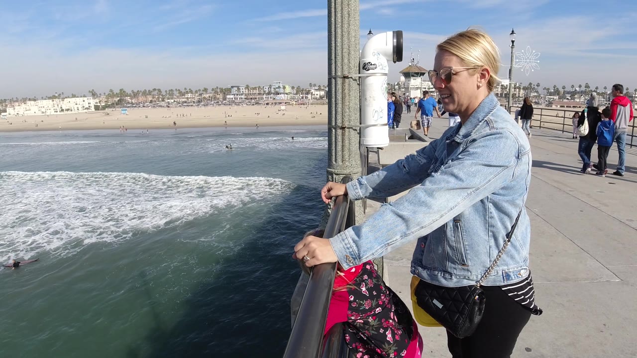 At 0:02 in Huntington Beach, California, the woman stands beside the railing with her hands forward, with darker water shadow below and a line of beach and distant people across the background.