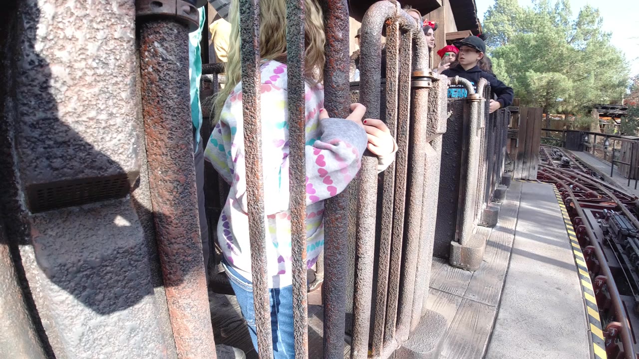 A child in a pastel patterned hoodie stands close behind rust-colored queue bars beside roller coaster tracks in Huntington Beach, California, with other riders visible farther down the line.