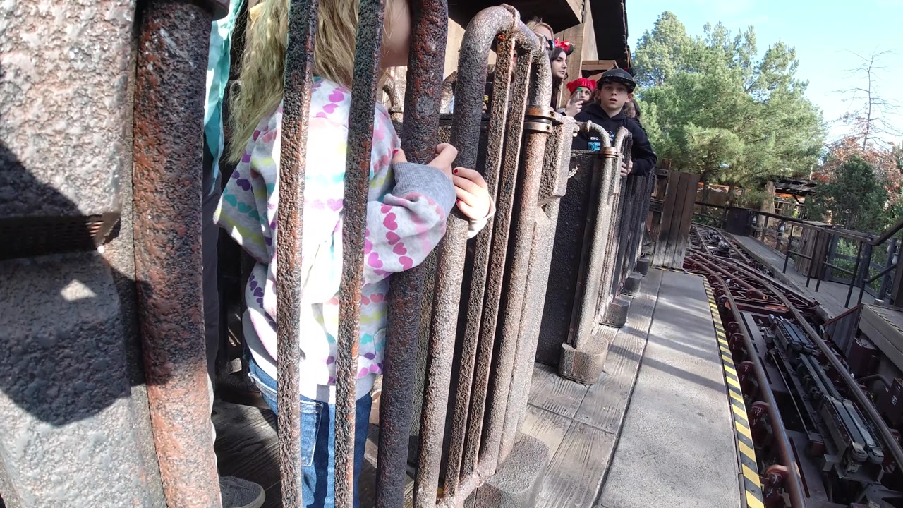 A close foreground of heavy queue bars partially blocks a child in a patterned hoodie, while roller coaster rails and waiting riders extend into the background in Huntington Beach, California.