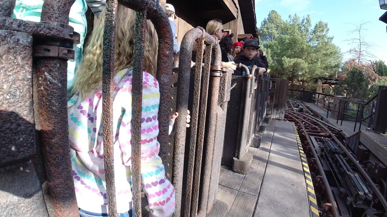 A young girl in a striped pastel shirt stands partly behind rust-textured metal gate bars near ride tracks in Huntington Beach, California.