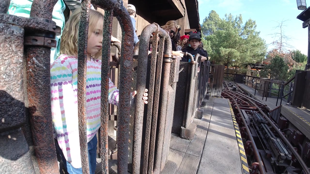 The girl faces the metal bars more directly, with her face visible between foreground posts and the ride track extending into the background in Huntington Beach, California.