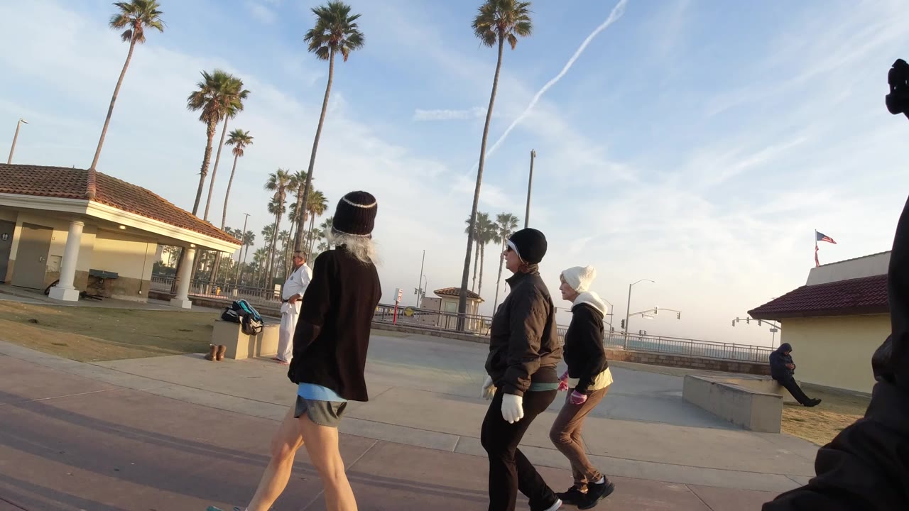 Teens walk across a Huntington Beach paved promenade, with tall palms, a tiled-roof public building, and soft daylight behind them.