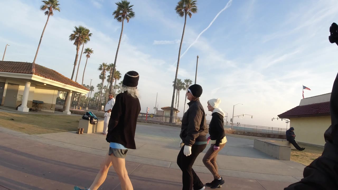 The group moves farther along the Huntington Beach walkway, with one walker in shorts closest to camera and the restroom-style building centered behind.