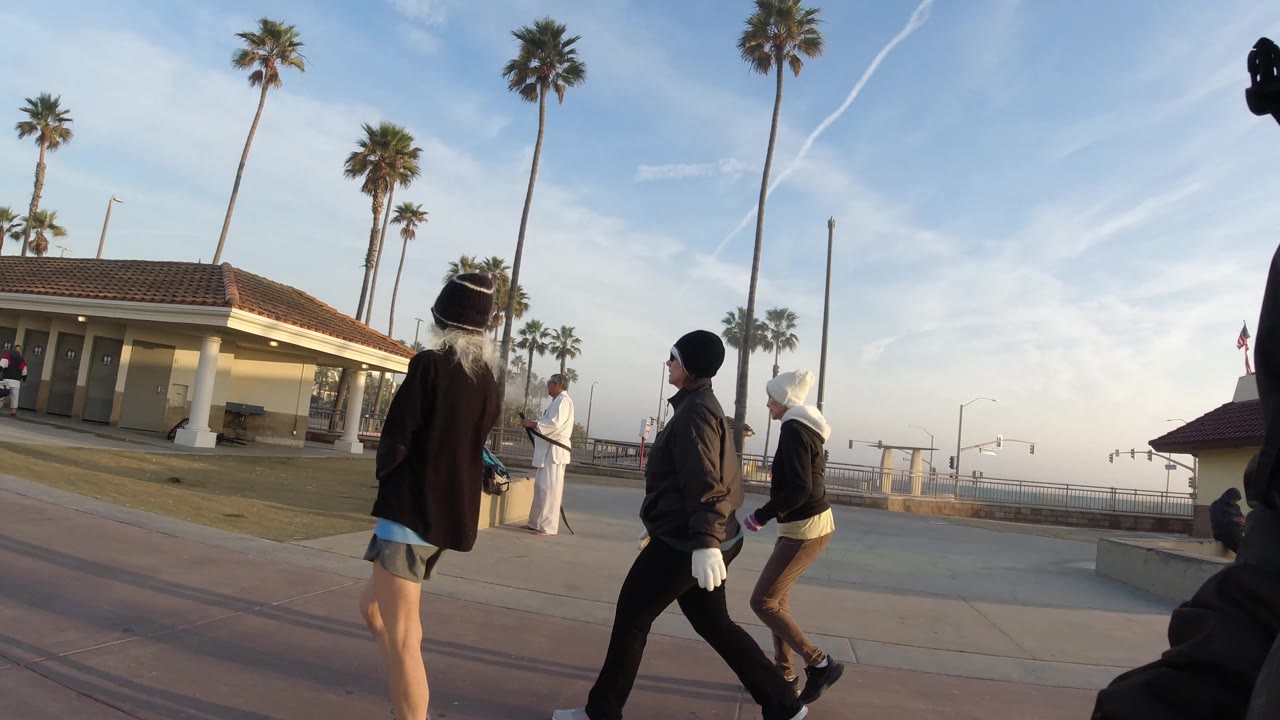 Three visible walkers cross the foreground of a Huntington Beach promenade while the low tiled-roof building, palms, and pale sky fill the background.