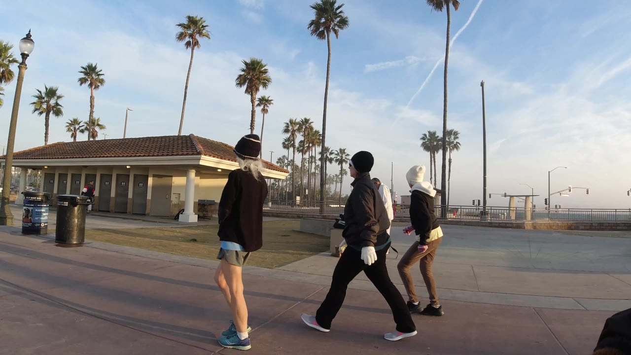 Teens walk near a public building, trash cans, palms, and a white vehicle.