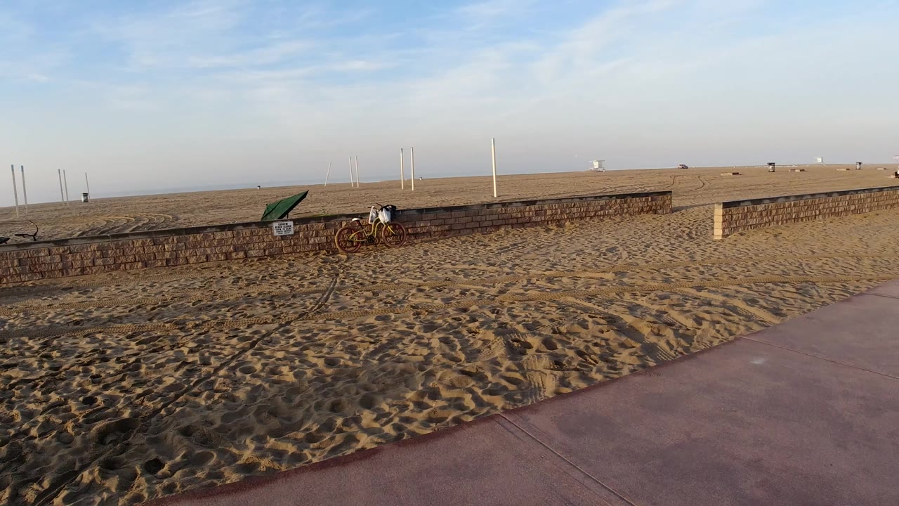 Wide view in Huntington Beach, California, showing rippled sand, a pink paved path, a low brick wall, bicycles, white poles, and a small person near the wall at the start of the clip.