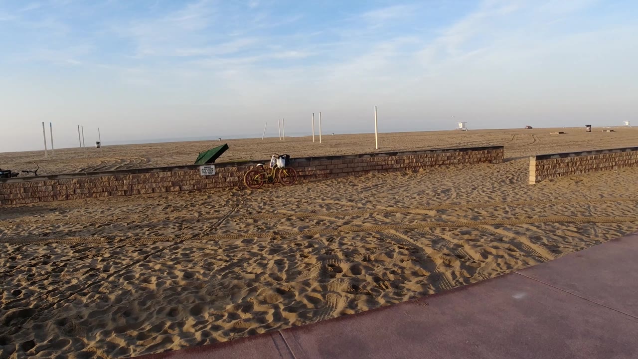 Huntington Beach, California, view with a broader sweep of sand and path, the low brick wall stretching across the middle, bicycles and a small person near the left side, and white poles on the horizon.