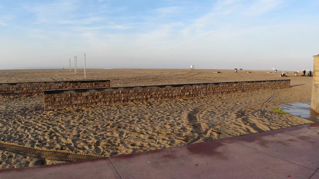 Wide Huntington Beach sand view from a sidewalk overlook, with a low seawall, red pavement, pale sky, and no visible people.