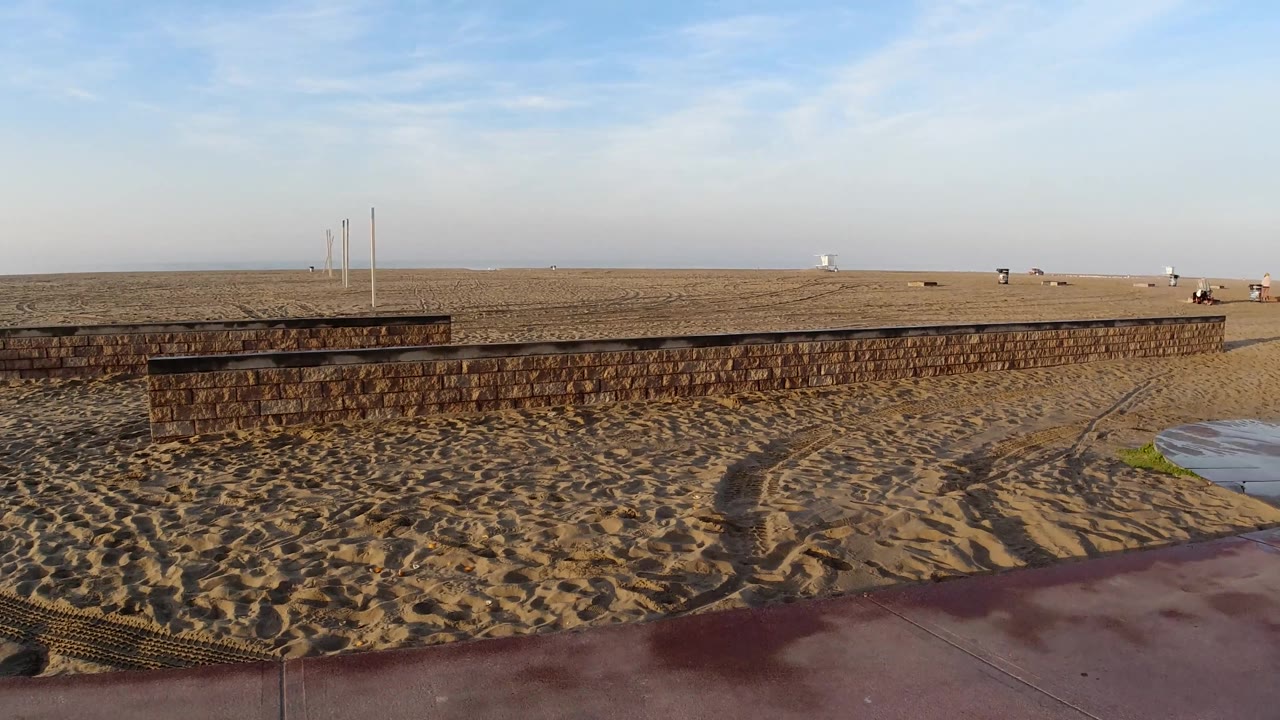 A broad Huntington Beach view shows rippled sand, a low wall across the midground, red sidewalk below, and open sky with no visible people.