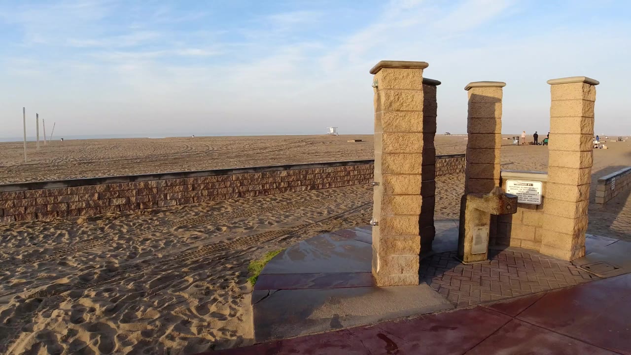 Stone pillars and a small fixture stand beside a sandy Huntington Beach walkway and low wall, with open beach and no clearly visible people in the near foreground.
