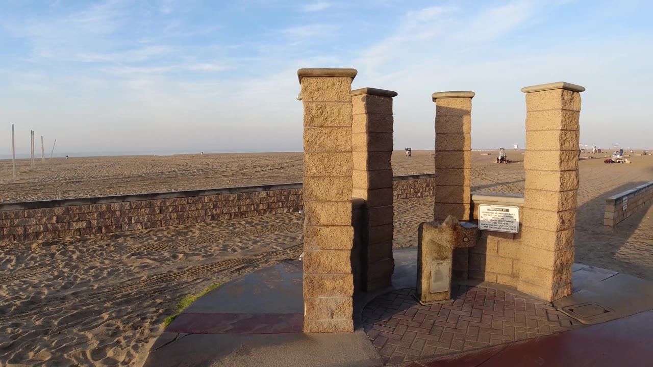 The pillar structure remains in the foreground at Huntington Beach while distant beachgoers and small structures appear across the open sand under a clear blue sky.