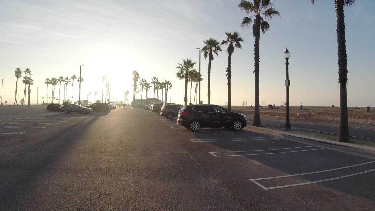 A Huntington Beach parking lot at sunrise with empty painted spaces in the foreground, parked cars at both sides, palm trees along the road, and small distant people near the beach.