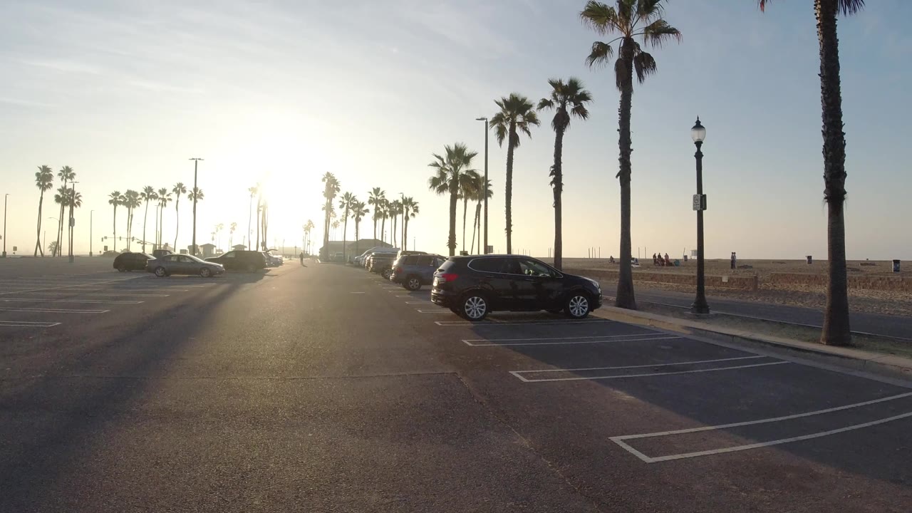At 0:01, the Huntington Beach lot remains wide and open, with parking lines leading toward a row of cars, tall palms, a bright sun near the horizon, and tiny people in the distance.