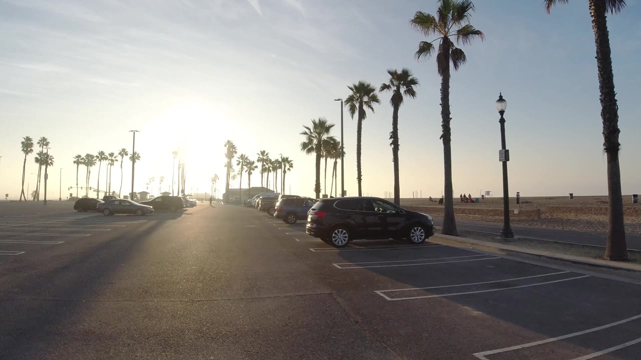 At 0:02, parked cars line the right side of the Huntington Beach parking area while tall palms frame the road and the low sun flares across the asphalt.