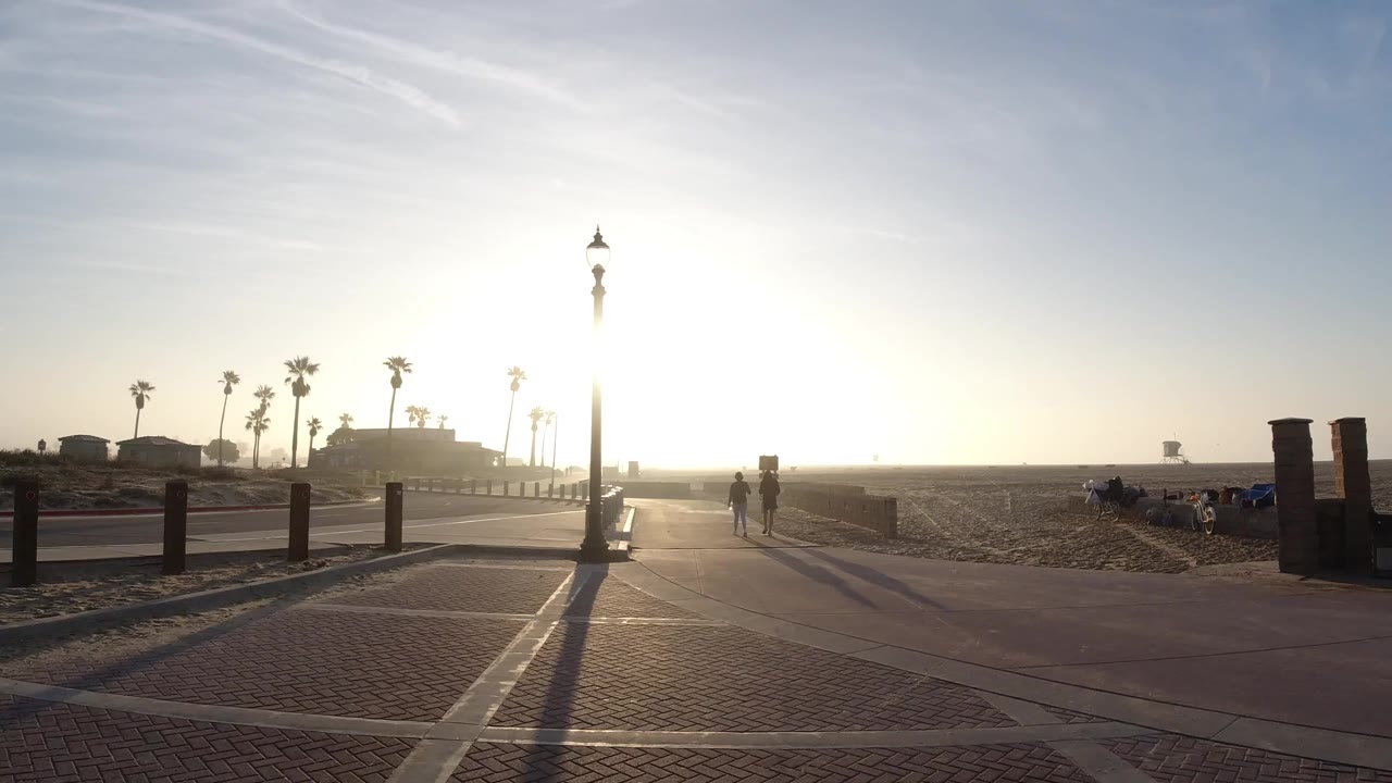 Wide backlit view of a Huntington Beach walkway with a central lamp post, brick paving, sand, palm trees, and small distant figures near the bright horizon.