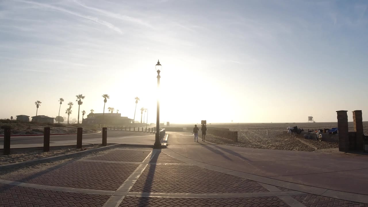 Sunlit beach path with lamp post, sand, palms, and pedestrians in the distance.