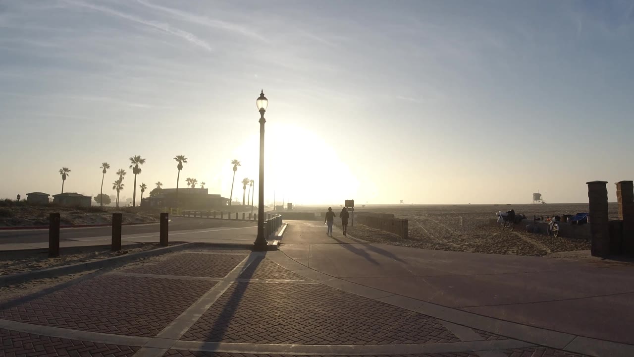 A Huntington Beach walkway curves past rail posts and sand as a lamp post stands against the sun, with people visible on the path and seated near the beach edge.
