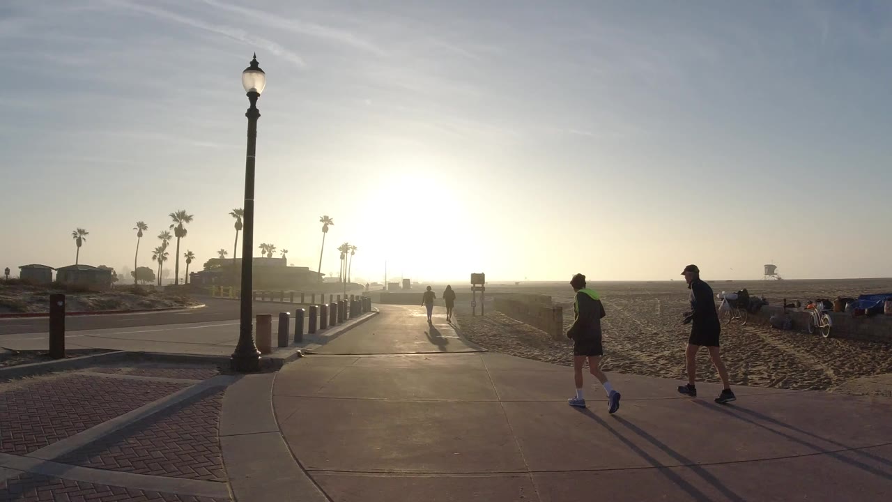 Backlit people walk and jog on a Huntington Beach path at sunrise, with a tall lamp post, sand, bollards, palms, and low buildings in view.