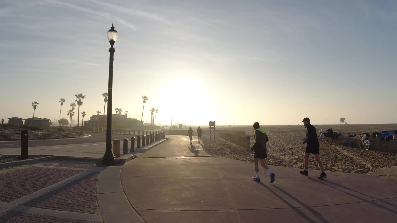 Two backlit people pass through the foreground on a Huntington Beach walkway, with sand, palm trees, bollards, and bright dawn glare behind them.