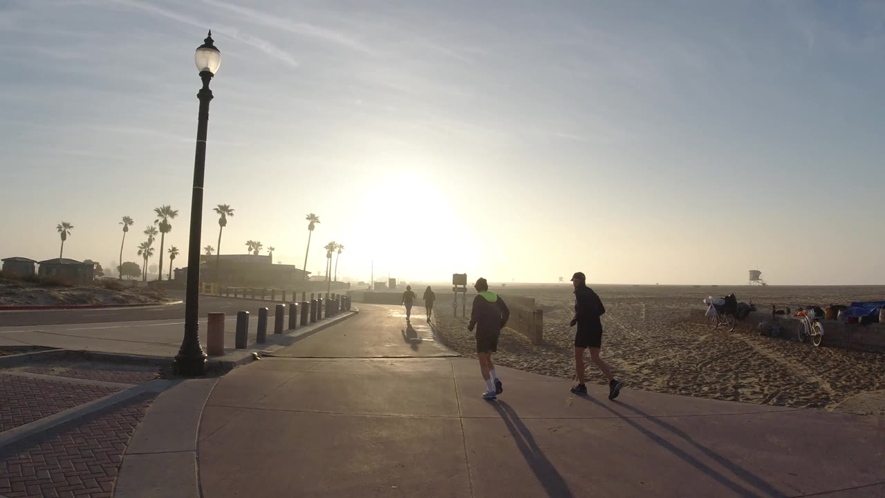 Several people move farther along the Huntington Beach path, with the sun low ahead, sand to the right, palms near the horizon, and long shadows stretching behind them.