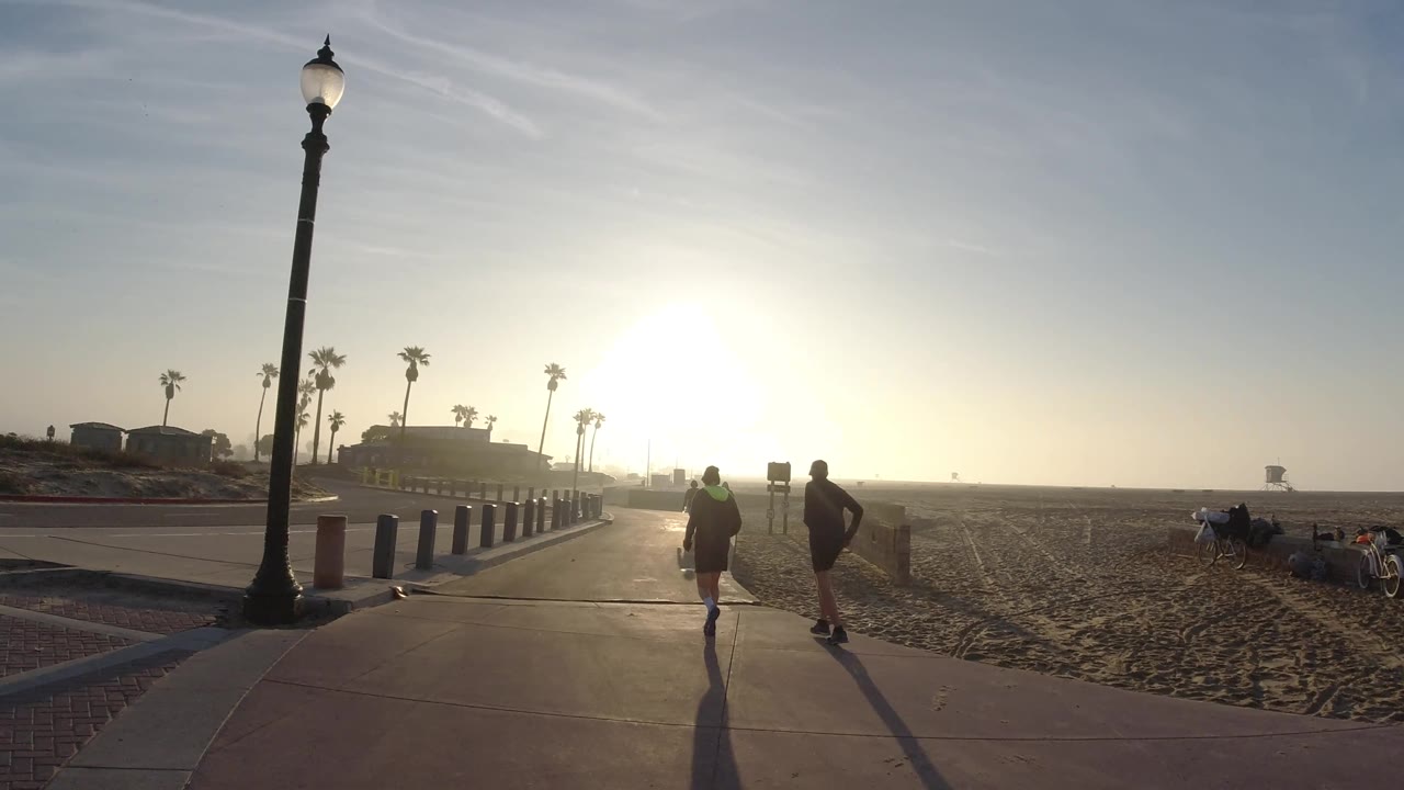 Backlit runner and walker on a beach path with sand and palms nearby.