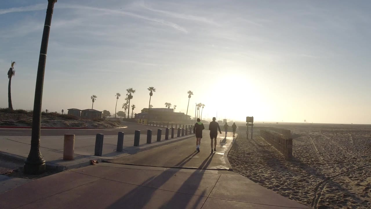At 0:00 in Huntington Beach, backlit pedestrians move along a sidewalk beside sand, blue bollards, palm trees, and low beachfront buildings.