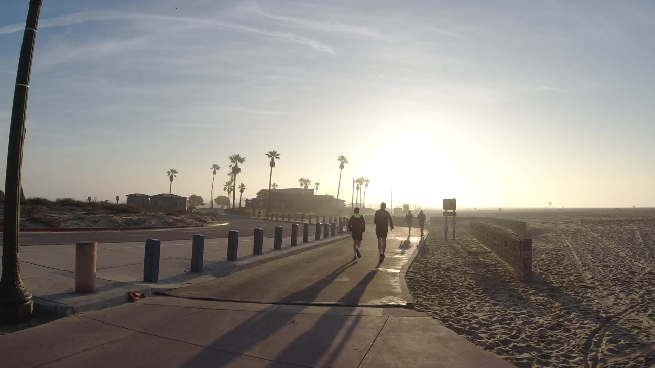 At 0:02 in Huntington Beach, silhouettes of walkers and a jogger pass the row of blue bollards while the sun sits low over the beach.