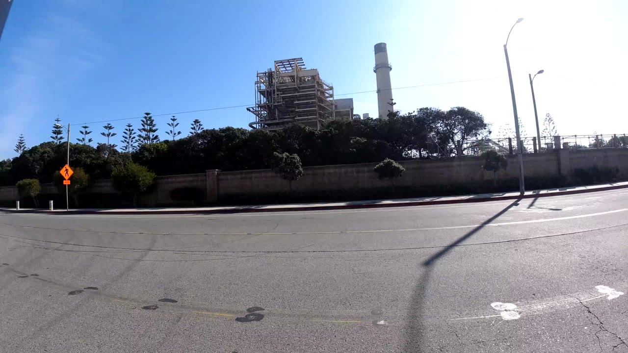 Sunlit road beside an industrial structure and tall stack.