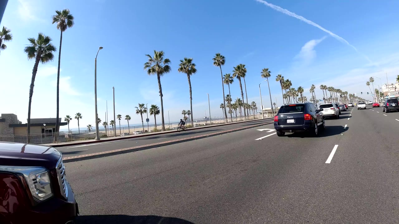 Wide Huntington Beach road view with moving cars, tall palm trees, bright blue sky, an ocean glimpse at left, and no clearly visible people.