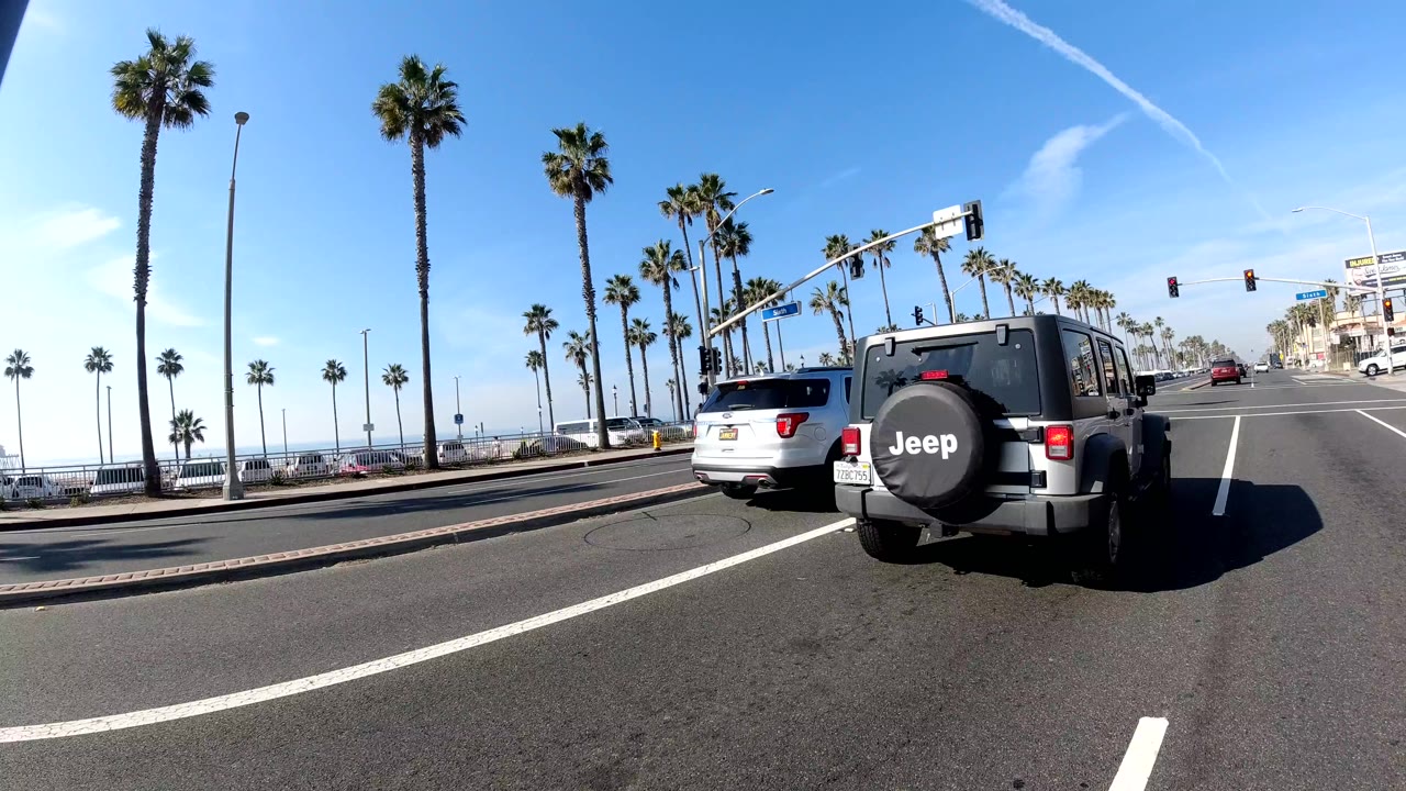 A Jeep and other vehicles approach a signalized Huntington Beach intersection with palm trees, traffic lights, bright sky, buildings at right, and no clearly visible people.