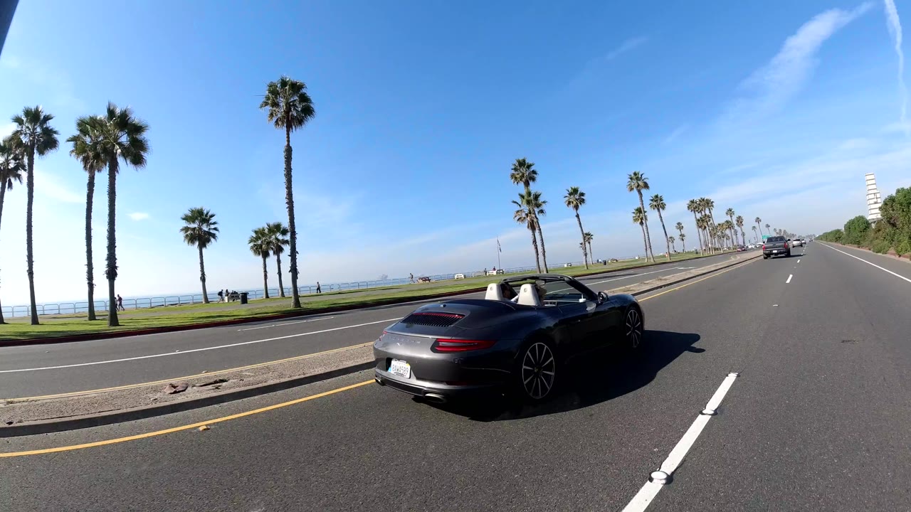 A dark convertible passes through a wide Huntington Beach roadway view with palm trees, coastline, lane markings, and no visible people.