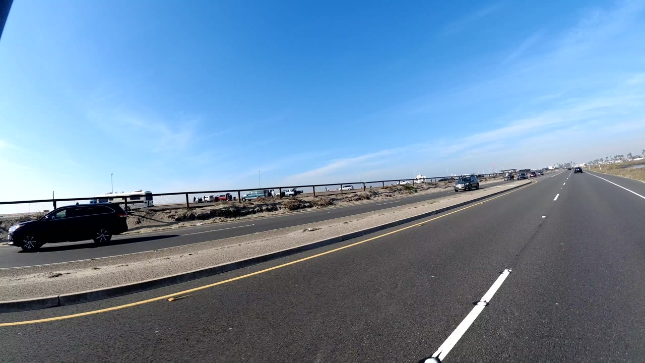 Wide Huntington Beach, California road view with lane markings, a black car at left, distant traffic, a low roadside barrier, bright blue sky, and no visible people.