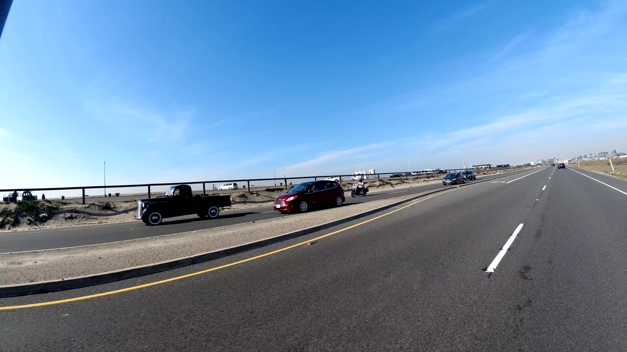 Huntington Beach, California roadway with a black off-road vehicle, a red car, open lanes, low roadside structures, blue sky, and no visible people.