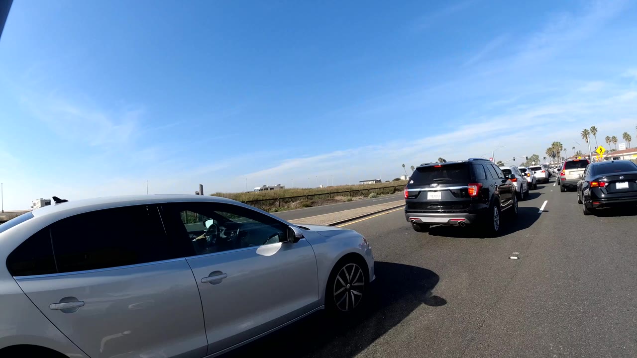 Huntington Beach, California traffic view with a white hatchback close to the camera, black SUVs ahead, palm trees, clear sky, and no visible people.