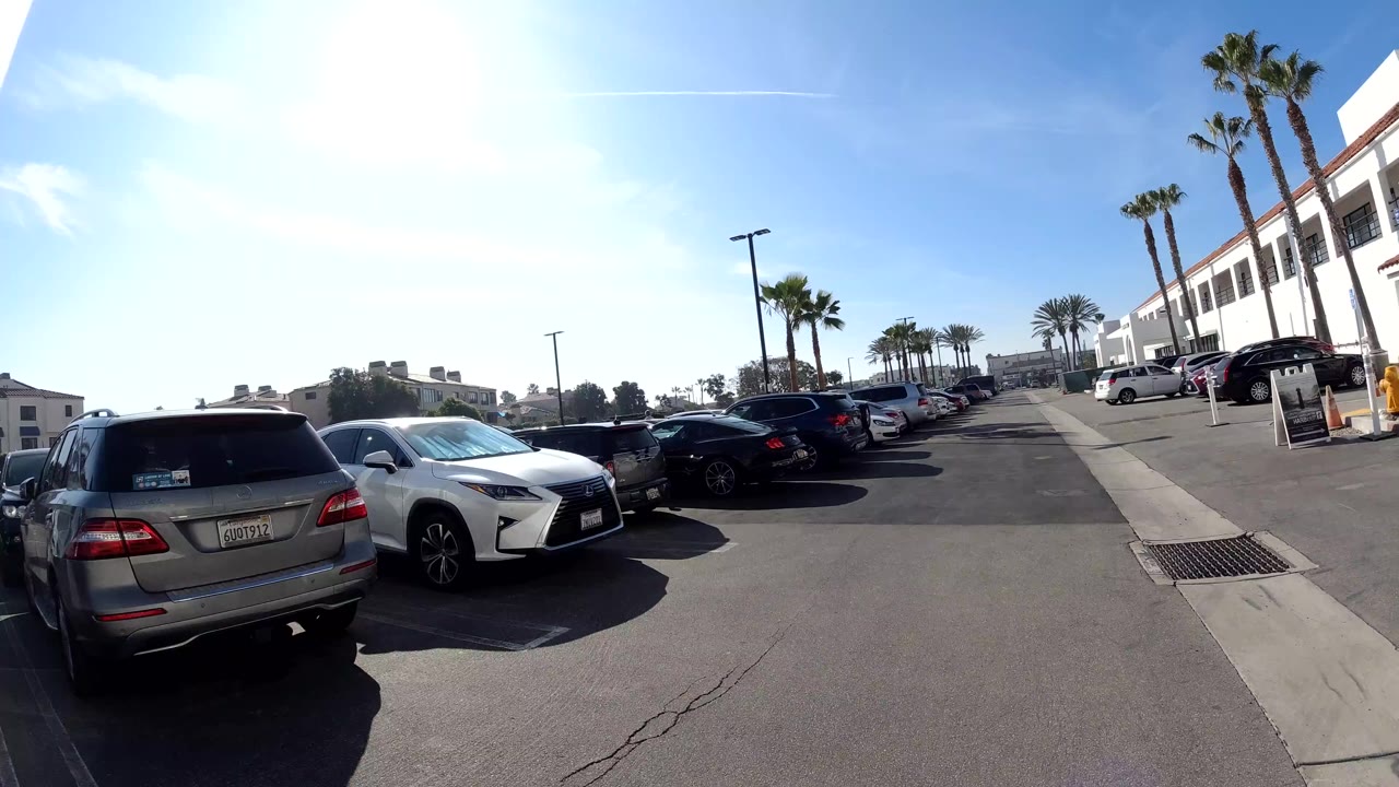Huntington Beach parking lot frame facing along the lane, with a gray SUV and white sedan in the foreground, palm trees and white commercial buildings to the right, strong sun glare, and no visible people.