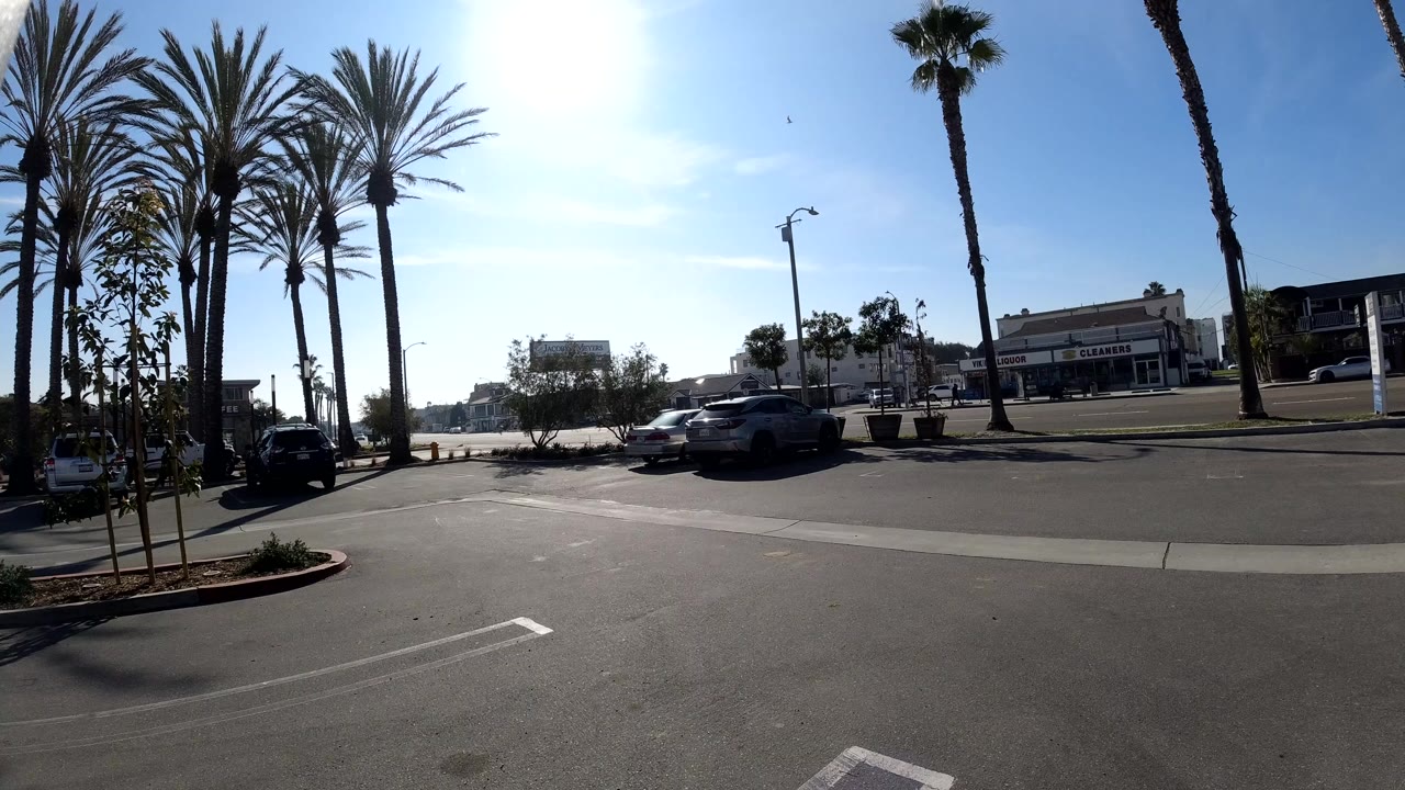 Opening frame of a sunny Huntington Beach parking lot with tall palms, parked cars, low buildings, bright blue sky, and no visible people.