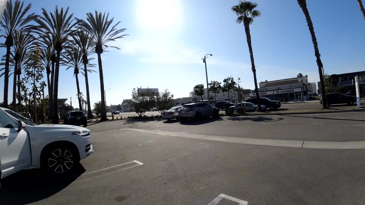 Huntington Beach parking-area view at the start of the clip, with a white car in the left foreground, palms overhead, storefronts across the road, and no visible people.